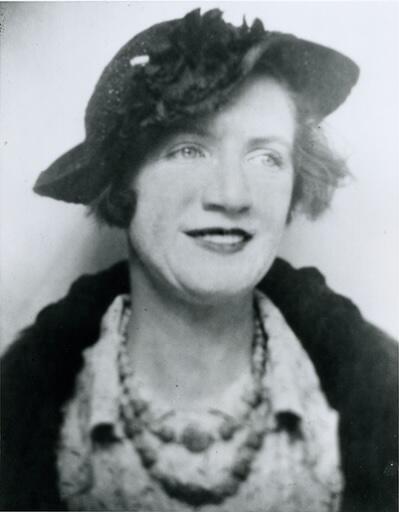 black and white photograph of a woman wearing a hat and a dark jacket and a necklance, looking off to the side with a slight smile on her face