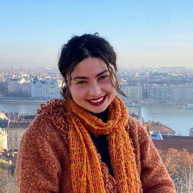 Aliza smiles at the camera. She is wearing an orange scarf and has a cityscape behind her.