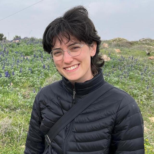 Maya Garfinkel, smiling, stands in front of a green field with small wildflowers and a grayblue sky. Maya has short dark hair, glasses, and a black puffer jacket.