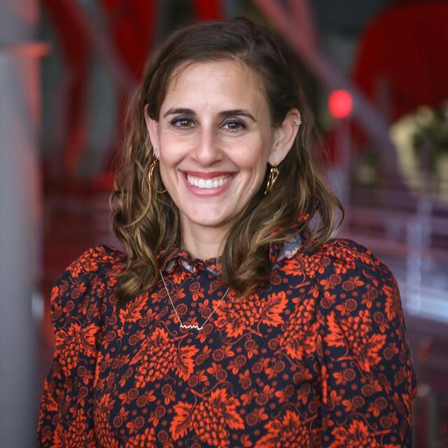 Headshot of Na'amit Sturm Nagel. She is a light skinned woman with brown eyes and wavy brown hair, smiling and wearing a red floral patterend shirt.