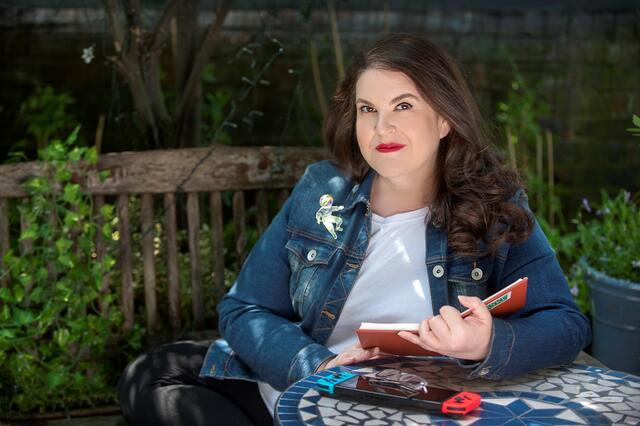 A portrait of Naomi Alderman, sitting on a bench and reading a book