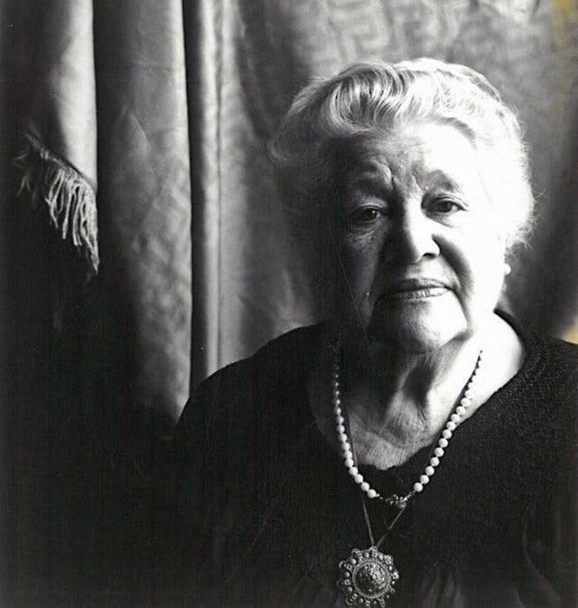 Black and white phtoograph of a woman with white hair wearing a dark dress looking pensively at the camera
