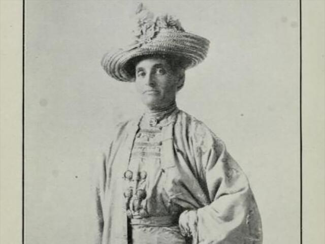Black and white historical photo of a woman wearing a light colored dress and a large hat, with a serious expression