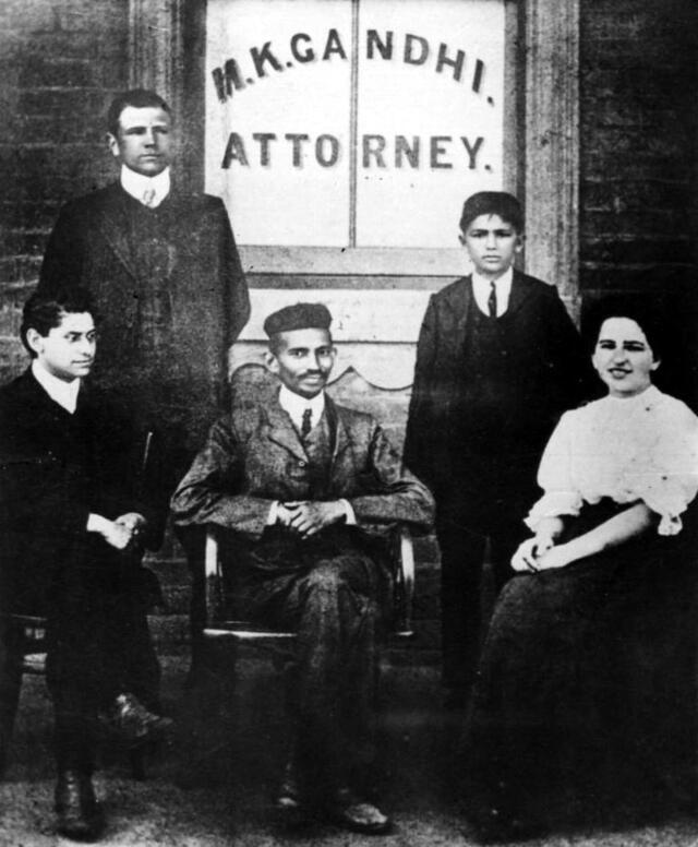 Sonja Schlesin with Gandhi and three others in front of a window reading M.K. Gandhi Attorney