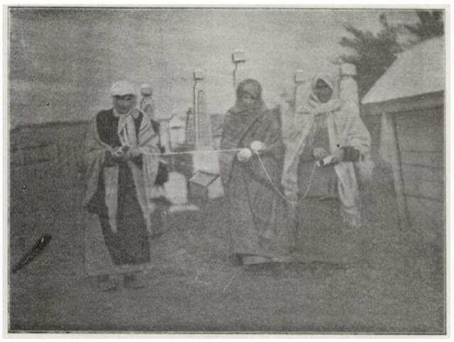 black and white historial photograph of women in robes and head scarves measuring a grave