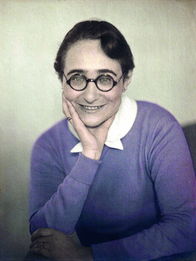 photograph of a seated woman wearing a purple sweater and round glasses, with her chin in her hand, smiling at the camera